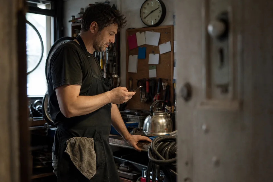 A service business owner in a workshop back room approves a draft on his phone, pinned pages on the cork board behind him